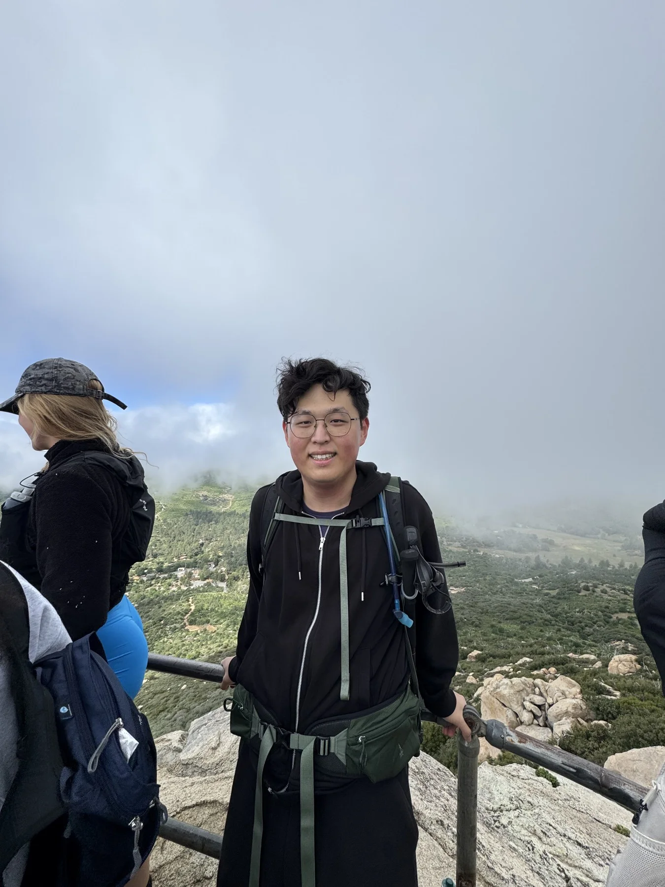 Hejia wearing a hiking backpack at a rocky overlook with low clouds behind him